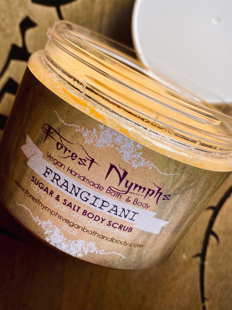 Puede incluir: Un tarro transparente de exfoliante corporal de az&uacute;car y sal de Frangipani con tapa blanca. La etiqueta dice "Forest Nymphs Vegan Handmade Bath & Body". El tarro contiene un exfoliante amarillo-anaranjado. La etiqueta es marr&oacute;n con texto blanco y morado.
