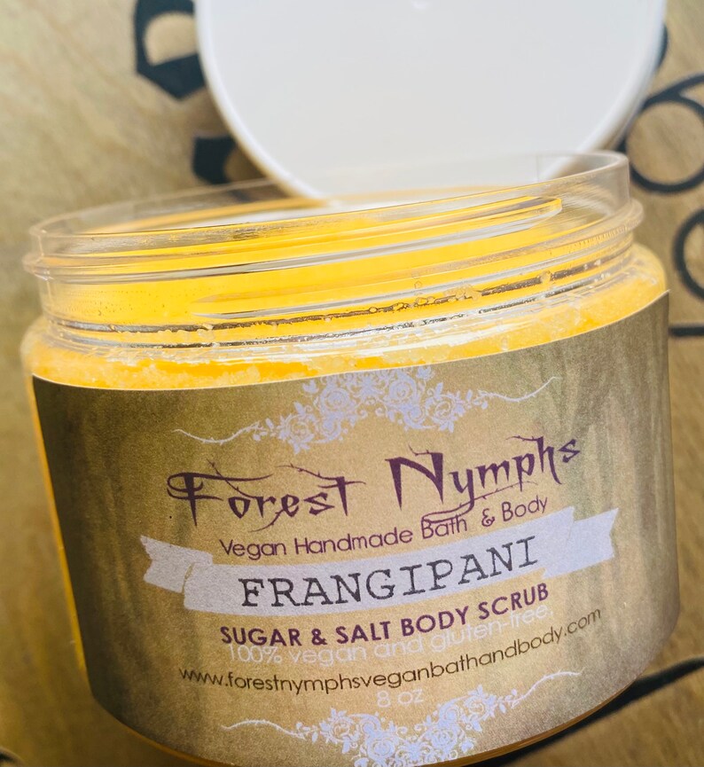 Puede incluir: Un tarro transparente de exfoliante corporal de az&uacute;car y sal Frangipani con tapa blanca. La etiqueta dice "Forest Nymphs" y "Vegan Handmade Bath & Body". El tarro contiene 227 gramos de producto.