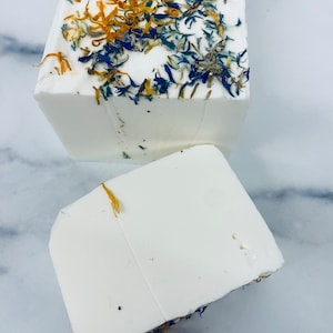 May include: Two rectangular bars of white soap, one whole and one cut, are displayed. The top of the soap is decorated with dried blue and orange flower petals. The soap is on a white and gray marble surface.
