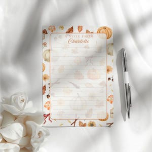 May include: A notepad with a fall theme, featuring pumpkins, leaves, and floral accents. The notepad has the text "A note from Charlotte" at the top. A silver pen with a white grip rests beside the notepad.