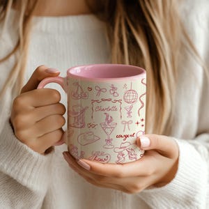 May include: A pink and beige ceramic mug with a pink interior, decorated with a repeating pattern of cowboy-themed illustrations in pink. The mug has the name "Charlotte" and the word "cowgirl" written on it.