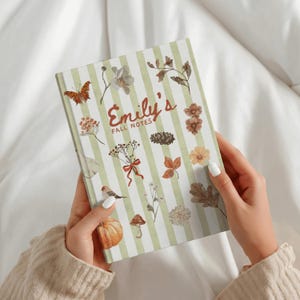 Pode incluir: Um caderno de capa dura com riscas verticais verde-claras e brancas, com ilustrações em aguarela de uma borboleta, flores, folhas, uma pinha, um pássaro, um cogumelo e uma abóbora, com o texto Emily's FALL NOTES.