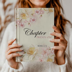 May include: A hardcover book with a floral design featuring daffodils and pink blossoms. The cover reads "Chapter Twenty One" with the name "Rebecca" below. The book has a cream-colored background.