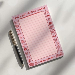May include: A pink notepad with the text "just a girl who loves cooking" and a spiral binding. The notepad is lined and has red illustrations of cooking-related items around the border. A black and silver pen rests beside the notepad.