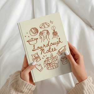 Könnte beinhalten: Ein cremefarbenes Notizbuch mit dem Titel "Sourdough Notes" mit braunen Illustrationen von Werkzeugen und Zutaten zum Brotbacken. Das Notizbuch wird von einer Person mit weißem Nagellack gehalten. Das Cover zeigt ein Brot, einen Mehlbeutel und eine Teigrolle.