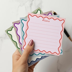 以下が含まれることがあります： 波状の縁取りが付いたカラフルなメモ帳の束を手に持っています。一番上のメモ帳はピンク色で、「Alana」という名前と罫線が書かれています。他のメモ帳は緑、紫、青、グレーです。金色のアクセントが付いた黒いペンが近くにあります。