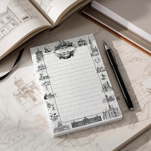 May include: A lined notepad with a decorative border of architectural drawings. The notepad is labelled "MICHELLE" at the top. A black pen lies next to the notepad, with an open book visible in the background.