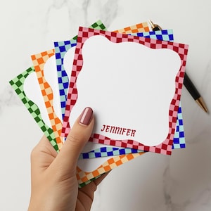 May include: A stack of personalized notepads with a white interior and a colorful checkered border in green, orange, blue, and red. The name "JENNIFER" is printed in red on the bottom of the notepad.