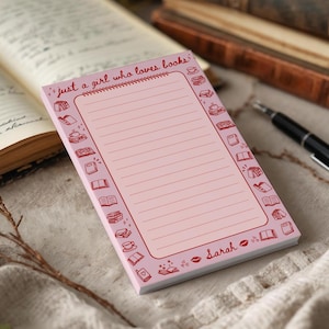 May include: A pink notepad with the text "just a girl who loves books" in red script. The notepad has a spiral binding and lined pages. Red illustrations of books and teacups border the notepad. The name "Sarah" is written in red script.