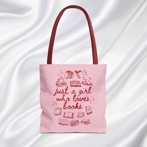 May include: Pink tote bag with a maroon handle featuring the text "just a girl who loves books" and illustrations of books, hearts, and a teacup. The bag is made of a soft material.