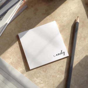 May include: A white square sticky note pad with the name "emily" written in cursive. A grey pencil lies next to the pad on a light brown surface. Other stationery items are visible in the background.