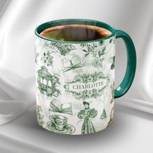 May include: White ceramic mug with a green handle and rim, filled with coffee. The mug features a green and white floral and book pattern with the name "CHARLOTTE" in a decorative frame. A charming and elegant design.