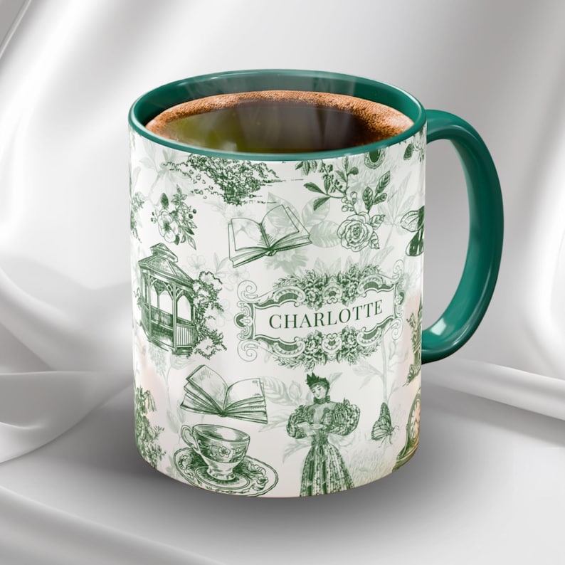 May include: White ceramic mug with a green handle and rim, filled with coffee. The mug features a green and white floral and book pattern with the name "CHARLOTTE" in a decorative frame. A charming and elegant design.