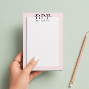 May include: A pink notepad with a black border and the text "DPT Doctor of Physical Therapy" and "Laura James" in black. A wooden pencil with a pink eraser rests on a light green surface.