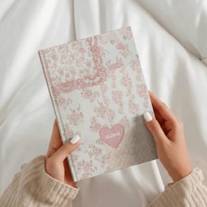 Könnte beinhalten: Ein Notizbuch mit festem Einband und einem hellen Blumenmuster in Rosa- und Weißtönen. Der Einband enthält unten in der Mitte einen dekorativen, herzförmigen Aufnäher in Rosa, auf dem der Name OLIVIA in weißen Großbuchstaben aufgedruckt ist.