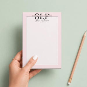 May include: A pink notepad with the text "SLP Speech-Language Pathologist" and the name "Laura James" in black. A wooden pencil with a pink eraser rests on a light green surface. The notepad is held by a hand.