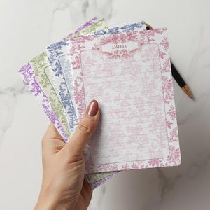 Op de afbeelding: Een hand houdt een stapel gelijnde notitieblokken met bloempatronen in verschillende kleuren vast. Het bovenste notitieblok is roze met de naam "AMELIA" bovenaan. Een zwarte pen met een gouden punt ligt op het oppervlak.