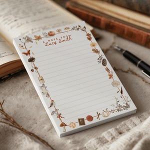 Può includere: Un blocco note rettangolare con linee orizzontali e un bordo decorativo con illustrazioni ad acquerello di zucche, funghi, fiori, foglie e uccelli. Il testo A NOTE FROM Lucy Smith è stampato in marrone nella parte superiore centrale.