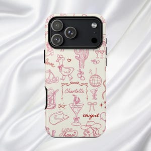 May include: A phone case with a cream background and a pink doodle design. The design includes a cowboy hat, cactus, martini glass, disco ball, and the word "cowgirl". The name "Charlotte" is also featured.