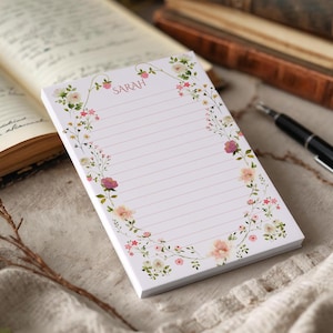 Puede incluir: Un bloc de notas blanco con páginas rayadas rosas y el nombre "SARAH" en la parte superior. El bloc de notas está decorado con una corona floral de flores rosas y blancas. Un bolígrafo y un libro abierto están en el fondo.