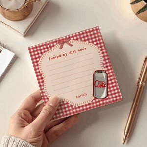 May include: A square notepad with a red and white gingham pattern featuring a central cream-colored scalloped label. The label includes a small red bow graphic, the text fueled by diet coke, a Diet Coke can illustration, and the name sarah.