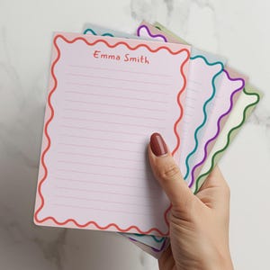 May include: A hand holds a stack of lined notepads with scalloped edges. The top notepad is pink with a red border and the name "Emma Smith" printed in the upper centre. Other notepads have teal, purple, and green borders.