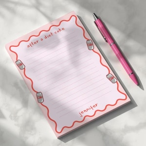 Peut inclure: Bloc-notes rose avec des pages lignées, une bordure ondulée rouge et des illustrations de canettes de Coca-Cola Light. Le bloc-notes porte l'inscription "after a diet coke" et "Jennifer". Un stylo rose est posé à côté.