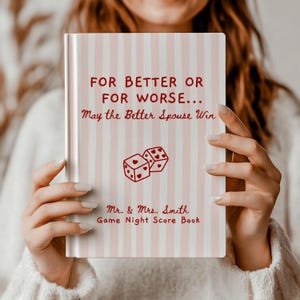 Puede incluir: Un libro de tapa dura con una cubierta de rayas verticales en rosa claro y blanco. El texto dice: FOR BETTER OR FOR WORSE... May the Better Spouse Win. Debajo hay un gráfico de dos dados con corazones, seguido de: Mr. & Mrs. Smith Game Night Score Book.