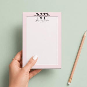 May include: A pink notepad with the text "NURSE PRACTITIONER" and "Laura James" in black. A wooden pencil rests on a light green surface next to the notepad. The notepad is held by a hand with a gold ring.