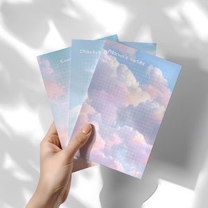 May include: Three notebooks with cloud-themed covers are held in a hand. The notebooks have a grid pattern and the text "Alana's notes", "Charlotte", and "Em". The covers feature a pastel sky with pink, blue, and white clouds.