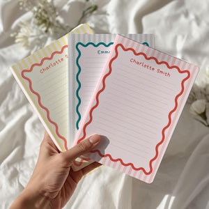 May include: Three personalised notepads with scalloped borders and striped backgrounds are held in a hand. The notepads are in pastel colours: yellow, green, and pink. Each notepad has a name printed at the top: "Charlotte Smith" and "Emma".