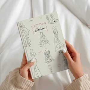 Pode incluir: Um diário de capa dura em tom claro com ilustrações de traço preto de vários vestidos, sapatos de salto alto e pequenos corações. O nome Jillian está impresso em letra cursiva preta no centro, emoldurado por um laço rosa desenhado.