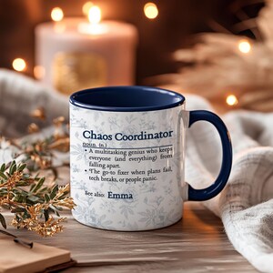 以下が含まれることがあります： 青い内側とハンドルが付いた白いセラミックマグ。マグには「Chaos Coordinator」のテキストと定義、そして「Emma」という名前が記載されています。マグには花柄があります。
