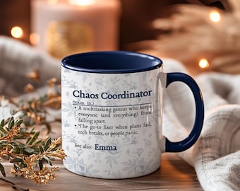 以下が含まれることがあります： 青い内側とハンドルが付いた白いセラミックマグ。マグには「Chaos Coordinator」のテキストと定義、そして「Emma」という名前が記載されています。マグには花柄があります。