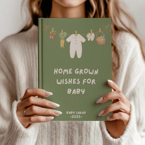 May include: A green journal with the text "HOME GROWN WISHES FOR BABY" and "BABY SARAH -2025-". The cover features illustrations of baby clothes, carrots, and a basket. The journal is held by a person.