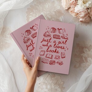 May include: Two pink journals with the text "just a girl who loves books" in red script. The cover design includes illustrations of books, hearts, and coffee cups. The journals are on a white surface with flowers.