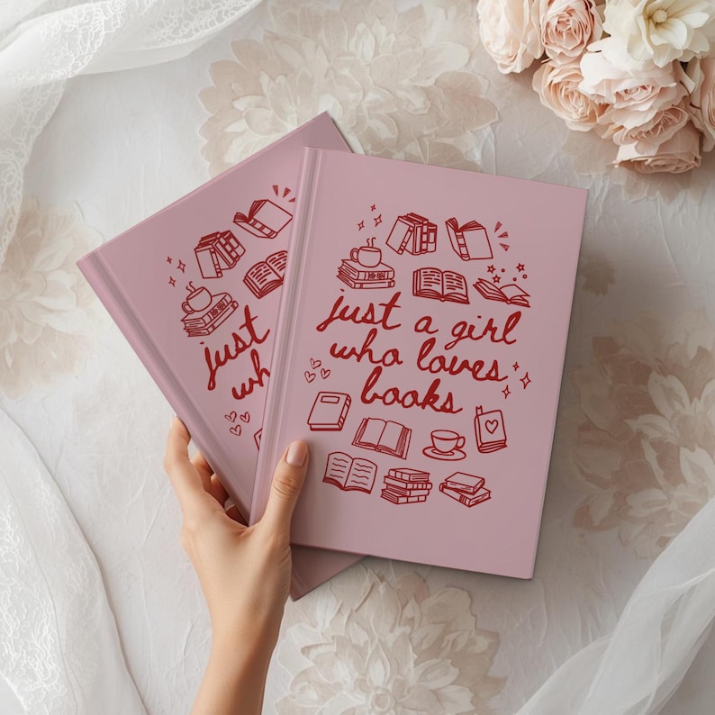 May include: Two pink journals with the text "just a girl who loves books" in red script. The cover design includes illustrations of books, hearts, and coffee cups. The journals are on a white surface with flowers.