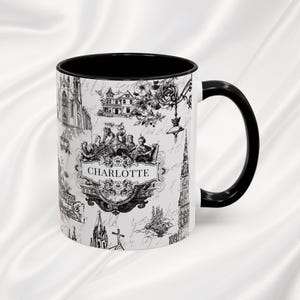 Könnte beinhalten: Weißer Keramikbecher mit schwarzem Innenraum und Griff. Der Becher zeigt eine Schwarz-Weiß-Illustration von Charlotte mit architektonischen und floralen Elementen. Das Wort "Charlotte" ist deutlich sichtbar.