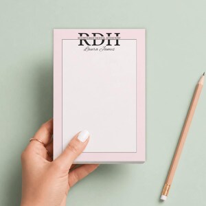 May include: A rectangular notepad with a light pink border and a white center. The top features the black text RDH, the words REGISTERED DENTAL HYGIENIST, and the name Laura James in a script font. A thin black line frames the white writing area.
