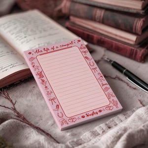 May include: A pink notepad with lined pages and a decorative border featuring cowboy-themed illustrations. The text "the life of a cowgirl" is printed at the top, with the name "Hayley" at the bottom. A pen and books are in the background.