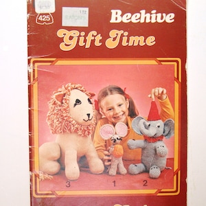 Op de afbeelding: Een vintage breipatronenboekje met de titel "Beehive Gift Time" met een rode en bruine omslag. De omslag toont een meisje dat een gehaakt olifantje, een leeuw en een muis vasthoudt. De tekst "Saison des Cadeaux" staat onderaan de omslag gedrukt.