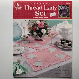 Può includere: Un libretto di modelli di uncinetto con un centrino in uncinetto rosa e bianco con un motivo floreale. Il libretto è intitolato "Thread Lady Set" e include un design originale di Annie's Attic. Il libretto include anche una foto di una mise en place con una tovaglia e sottopiatti in uncinetto rosa e bianco.