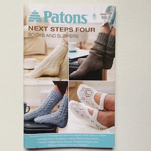 Peut inclure: Couverture du livre "Patons Next Steps Four Socks and Slippers" avec des images de chaussettes et de pantoufles tricotées de différents styles et couleurs. Le logo Patons et le code "5008 DD" sont également présents.