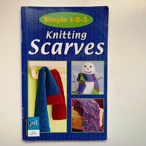 Peut inclure: Un livre intitulé "Knitting Scarves" avec une couverture bleue et des images d'écharpes tricotées et d'un bonhomme de neige. La couverture présente le texte "Simple 1-2-3" et "Knitting Scarves" en blanc. Le livre comprend des images d'écharpes bleues, rouges et à motifs.