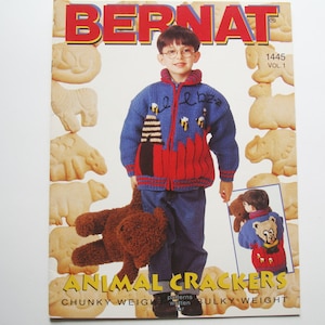 Puede incluir: Una portada de libro de patrones de tejido de punto con un niño que lleva un suéter de punto azul y rojo con galletas de animales. El título del libro es "Animal Crackers" y el texto "Chunky Weight patterns written for Bulky Weight yarn" está debajo del título.