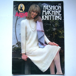 Op de afbeelding: Een tijdschriftcover met een vrouw die een witte gebreide jurk en een blauw-witte sjaal draagt. Het tijdschrift is getiteld "Argyll Fashion Machine Knitting" en het nummer 414 staat rechtsboven gedrukt.