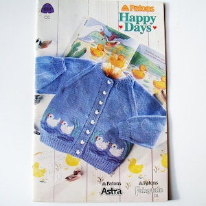 Könnte beinhalten: Blauer Baby-Strickcardigan mit weißen Knöpfen und Entenküken-Stickerei. Der Cardigan wird auf einem Vintage-Strickmusterbuch von Patons mit dem Titel "Happy Days" präsentiert. Das Buch enthält Illustrationen von Enten.