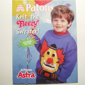 May include: A blue knitted sweater with a clown face design. The sweater is advertised as "Knit the Fleecy Sweater!" and is a Patons Astra product.