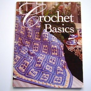 May include: Crochet Basics book cover featuring a purple and white crocheted blanket with a floral design. The book cover also features a cup of tea and a doily.