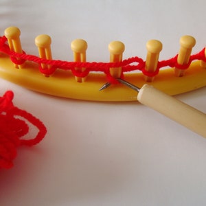 Peut inclure: Gros plan sur un fil rouge utilisé pour tricoter sur un métier à tisser circulaire jaune. Une aiguille à tricoter en bois est utilisée pour tirer le fil à travers les boucles.